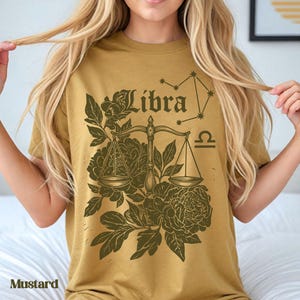 Op de afbeelding: Mosterdkleurig T-shirt met een Weegschaal sterrenbeeld ontwerp. Het ontwerp bevat het woord "Libra", een weegschaal, bloemelementen en een sterrenbeeld. Het shirt is gemaakt van een zacht materiaal.