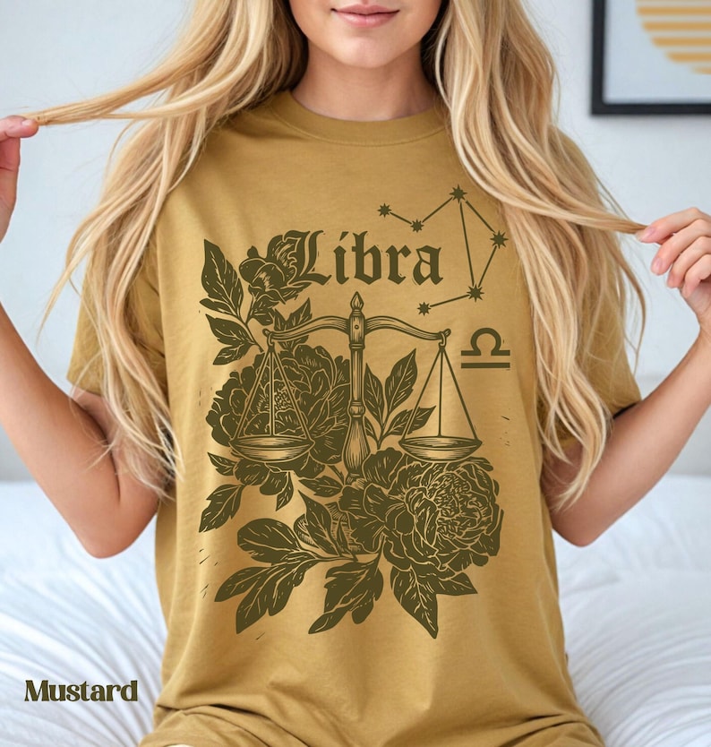 Op de afbeelding: Mosterdkleurig T-shirt met een Weegschaal sterrenbeeld ontwerp. Het ontwerp bevat het woord "Libra", een weegschaal, bloemelementen en een sterrenbeeld. Het shirt is gemaakt van een zacht materiaal.