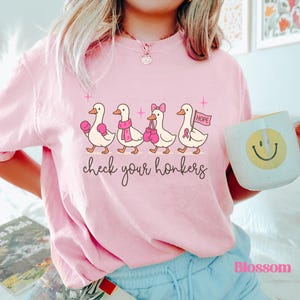 Pu&ograve; includere: T-shirt rosa chiaro con un motivo di anatre dei cartoni animati. Le anatre indossano guantoni da boxe, sciarpe e fiocchi. Il testo "check your honkers" &egrave; scritto sotto.