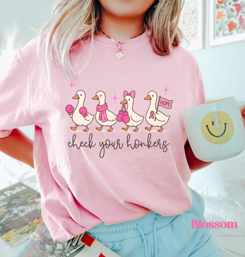 Pu&ograve; includere: T-shirt rosa chiaro con un motivo di anatre dei cartoni animati. Le anatre indossano guantoni da boxe, sciarpe e fiocchi. Il testo "check your honkers" &egrave; scritto sotto.