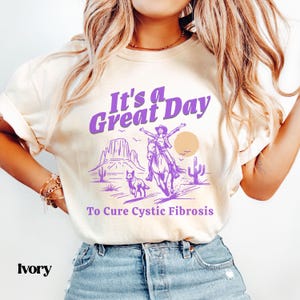 Puede incluir: Una mujer lleva una camiseta de color crema con texto morado que dice "It's a Great Day To Cure Cystic Fibrosis". El gráfico de la camiseta muestra a una mujer montando a caballo en un paisaje desértico.