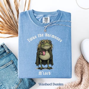 May include: A washed denim blue t-shirt featuring a graphic of a green frog with long hair wearing a dark cloak and holding a pink mug. The text above the frog reads Twas the Hormones and below it reads M'Lord. The shirt has a Comfort Colors label.