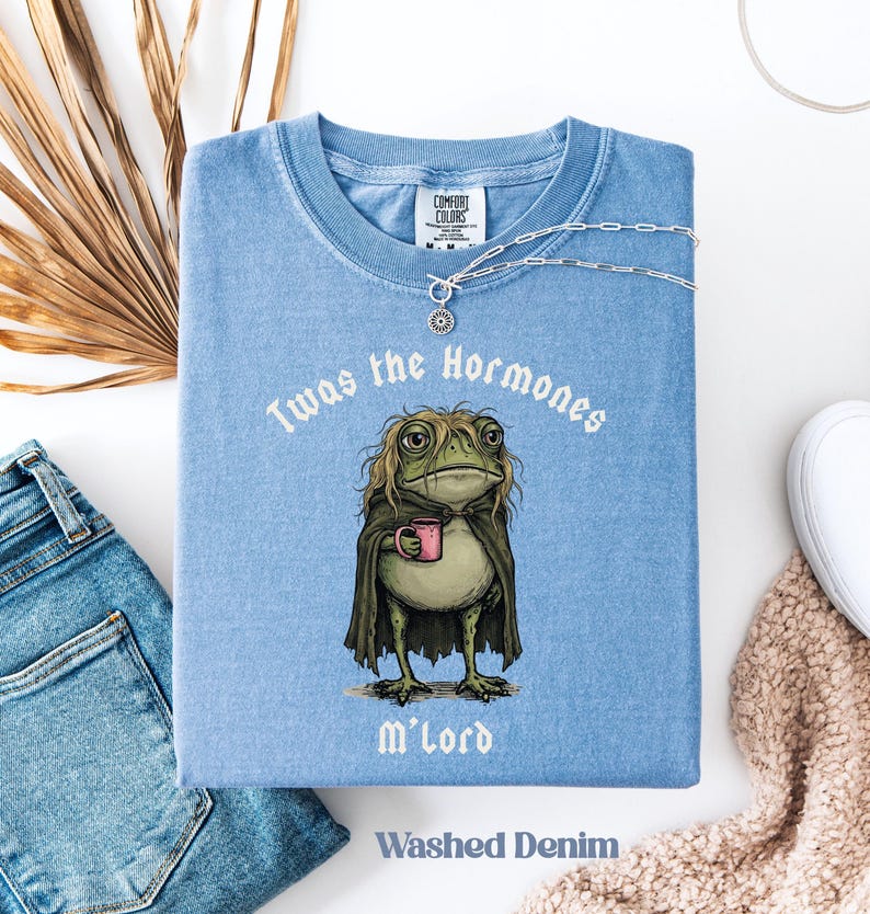May include: A washed denim blue t-shirt featuring a graphic of a green frog with long hair wearing a dark cloak and holding a pink mug. The text above the frog reads Twas the Hormones and below it reads M'Lord. The shirt has a Comfort Colors label.