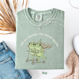 May include: A sage green t-shirt with the text "Off to change my mind again" encircling a graphic of two green frogs riding scooters. The shirt also features the word "Bay" at the bottom.