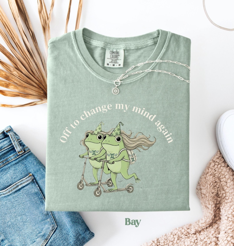 May include: A sage green t-shirt with the text "Off to change my mind again" encircling a graphic of two green frogs riding scooters. The shirt also features the word "Bay" at the bottom.