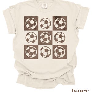 May include: Ivory t-shirt with a brown graphic of nine soccer balls arranged in a grid pattern.