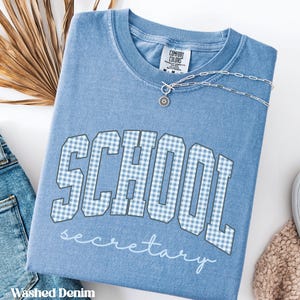 Op de afbeelding: Een t-shirt met ronde hals in gewassen denimblauw met het woord SCHOOL in een lichtblauw en wit geblokt lettertype, met daaronder het woord secretary in witte schrijfletters. Een zilveren ketting met een kleine ronde hanger ligt erbovenop.