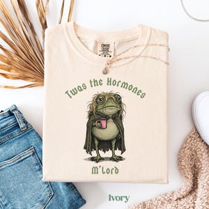 May include: An ivory t-shirt featuring a graphic of a green frog with messy hair wearing a dark cloak and holding a pink mug. The text above the frog reads Twas the Hormones and the text below reads M'lord. The shirt has a crew neckline.
