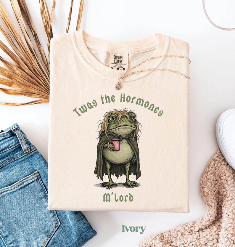 May include: An ivory t-shirt featuring a graphic of a green frog with messy hair wearing a dark cloak and holding a pink mug. The text above the frog reads Twas the Hormones and the text below reads M'lord. The shirt has a crew neckline.
