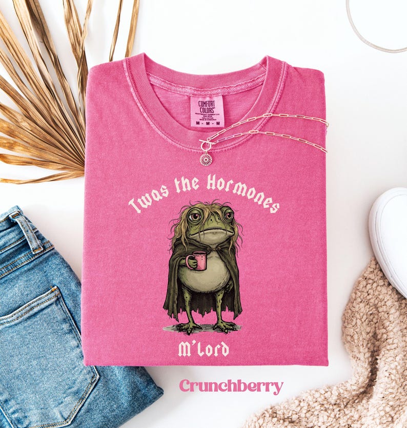 May include: A pink short-sleeved t-shirt featuring a graphic of a frog with long hair wearing a dark cloak and holding a pink mug. The text above the frog reads Twas the Hormones and the text below reads M'lord. The shirt has a crew neckline.