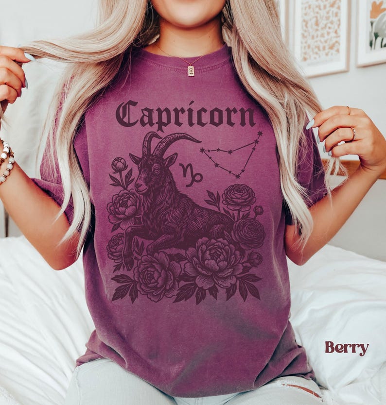 May include: Berry-colored t-shirt featuring a Capricorn design. The graphic includes the word "Capricorn" in a gothic font, a goat illustration, the Capricorn symbol, and a constellation. The design is surrounded by floral elements.
