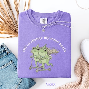 May include: A violet Comfort Colors sweatshirt with a graphic of two green frogs riding scooters, with the text "Off to change my mind again." A silver necklace with a sun pendant is draped on the shirt. The word "Violet" is printed at the bottom.
