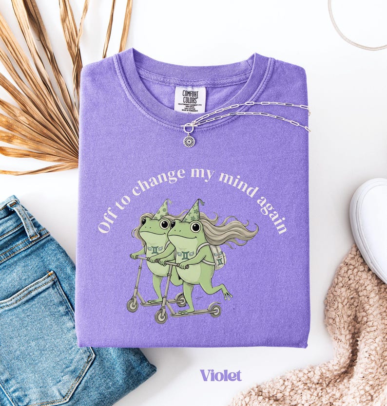 May include: A violet Comfort Colors sweatshirt with a graphic of two green frogs riding scooters, with the text "Off to change my mind again." A silver necklace with a sun pendant is draped on the shirt. The word "Violet" is printed at the bottom.