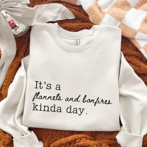 Puede incluir: Sudadera color crema con el texto negro "It's a flannels and bonfires kinda day". La palabra "Sand" est&aacute; impresa en la parte inferior. La sudadera se muestra sobre una superficie marr&oacute;n texturizada.