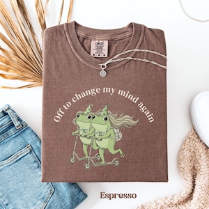 May include: Espresso-colored t-shirt with the phrase "Off to change my mind again" encircling a graphic of two green frogs riding scooters. The shirt has a crew neck and short sleeves. A silver necklace with a pendant is draped on the shirt.