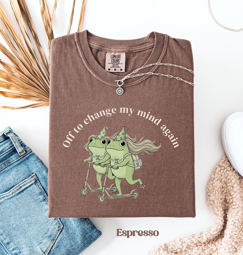 May include: Espresso-colored t-shirt with the phrase "Off to change my mind again" encircling a graphic of two green frogs riding scooters. The shirt has a crew neck and short sleeves. A silver necklace with a pendant is draped on the shirt.