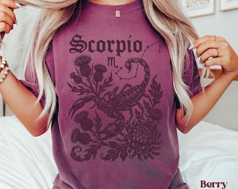 Op de afbeelding: Bessenkleurig T-shirt met een dierenriemontwerp van Schorpioen. De afbeelding bevat het woord "Scorpio", een schorpioen, een sterrenbeeld en bloemmotieven. Het shirt is een comfortabele, casual stijl.