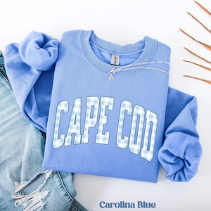 Op de afbeelding: Een Carolina blauwe sweatshirt met de tekst "CAPE COD" in een wit en lichtblauw ruitpatroon. Een zilveren ketting met een hanger ligt op de ronde hals. De sweatshirt is op een blauwe jeans gevouwen.