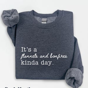 Puede incluir: Sudadera gris oscuro con el texto blanco "It's a flannels and bonfires kinda day". Las mangas est&aacute;n dobladas. La sudadera es de la marca Gildan.