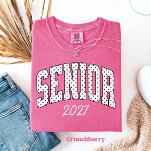 Peut inclure: Un t-shirt rose pli&eacute; avec le mot "SENIOR" en lettres &agrave; pois blancs et l'ann&eacute;e "2027" imprim&eacute;e sur le devant. Un collier argent&eacute; est drap&eacute; sur le t-shirt. Le t-shirt est sur une surface blanche avec un jean bleu et une chaussure blanche.