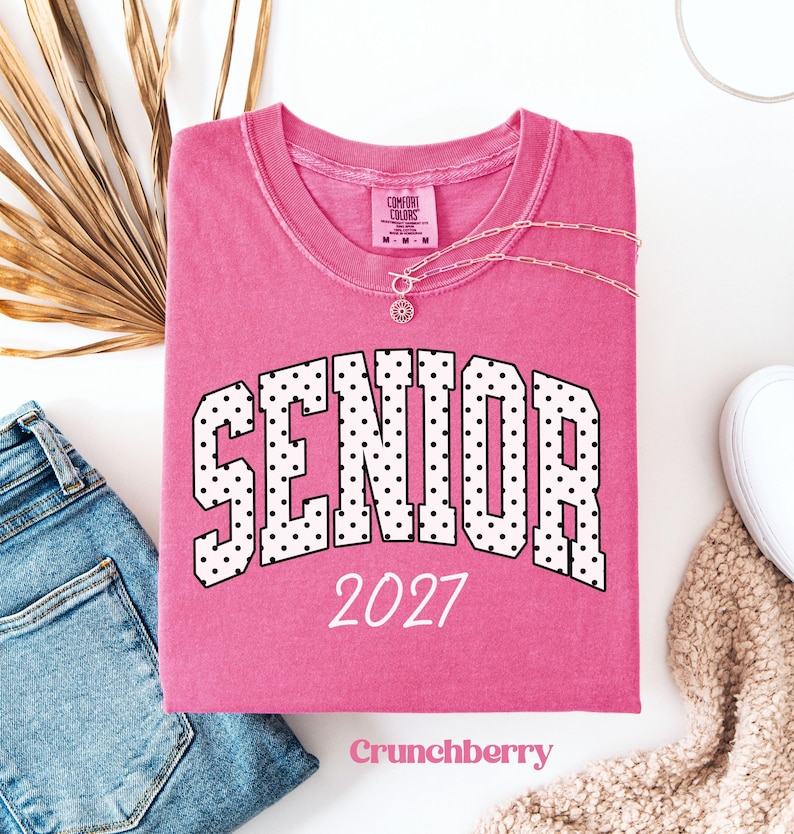 Peut inclure: Un t-shirt rose pli&eacute; avec le mot "SENIOR" en lettres &agrave; pois blancs et l'ann&eacute;e "2027" imprim&eacute;e sur le devant. Un collier argent&eacute; est drap&eacute; sur le t-shirt. Le t-shirt est sur une surface blanche avec un jean bleu et une chaussure blanche.