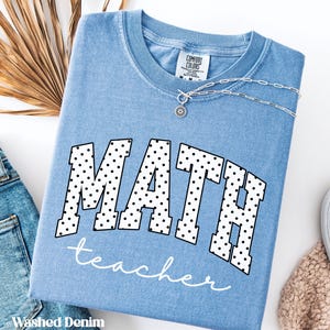 May include: A light blue "Washed Denim" t-shirt with the word "MATH" in large, black polka-dotted letters, and the word "teacher" in cursive below. A silver necklace with a pendant is visible.