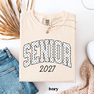 Peut inclure: T-shirt de couleur ivoire avec le mot "SENIOR" dans un motif &agrave; pois et l'ann&eacute;e "2027" imprim&eacute;e en noir. Le t-shirt est pli&eacute; sur une surface blanche &agrave; c&ocirc;t&eacute; d'un jean bleu et d'un pull beige.