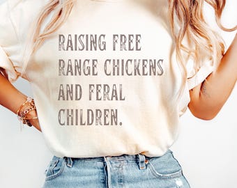 May include: A white t-shirt with the text "Raising Free Range Chickens and Feral Children." printed in brown on the front. The shirt is being worn by a person with long blonde hair and is styled with a gold necklace and bracelets.