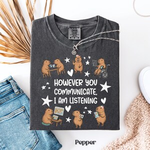 Op de afbeelding: Donkergrijs t-shirt met een afbeelding van capibara's die computers en tablets gebruiken, met de tekst "HOWEVER YOU COMMUNICATE, I AM LISTENING." Het shirt is van het merk Comfort Colors.
