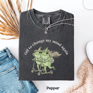 May include: A dark gray t-shirt with the text "Off to change my mind again" and a graphic of two green frogs riding scooters. The shirt has a Comfort Colors tag and a silver necklace. The word "Pepper" is printed at the bottom.