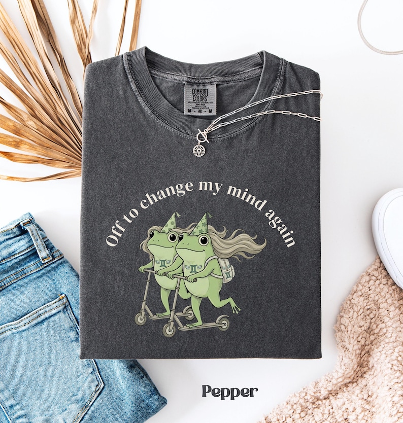 May include: A dark gray t-shirt with the text "Off to change my mind again" and a graphic of two green frogs riding scooters. The shirt has a Comfort Colors tag and a silver necklace. The word "Pepper" is printed at the bottom.