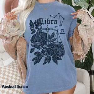 Op de afbeelding: Een blauw t-shirt met een Weegschaal sterrenbeeld ontwerp. Het ontwerp bevat het woord "Libra", een weegschaal, bloemelementen en een sterrenbeeld. Het shirt heeft ook de woorden "Washed Denim" aan de onderkant.