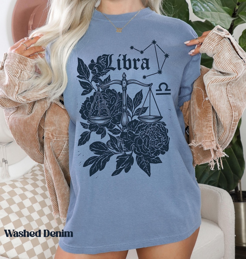 Op de afbeelding: Een blauw t-shirt met een Weegschaal sterrenbeeld ontwerp. Het ontwerp bevat het woord "Libra", een weegschaal, bloemelementen en een sterrenbeeld. Het shirt heeft ook de woorden "Washed Denim" aan de onderkant.