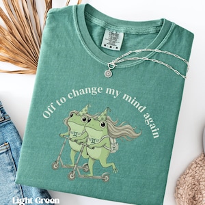 May include: Light green t-shirt with the text "Off to change my mind again" and a graphic of two frogs riding scooters. The shirt has a crew neck and a silver necklace with a sun pendant. The shirt is folded on a white surface.