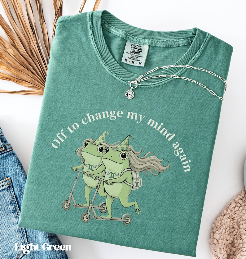 May include: Light green t-shirt with the text "Off to change my mind again" and a graphic of two frogs riding scooters. The shirt has a crew neck and a silver necklace with a sun pendant. The shirt is folded on a white surface.