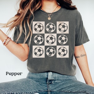 May include: A charcoal grey t-shirt with a graphic of nine white soccer balls arranged in a grid pattern. The soccer balls have a distressed look.