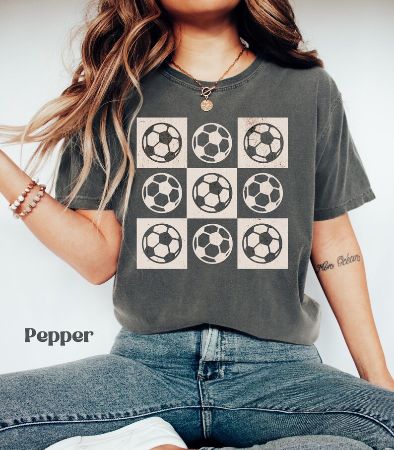 May include: A charcoal grey t-shirt with a graphic of nine white soccer balls arranged in a grid pattern. The soccer balls have a distressed look.