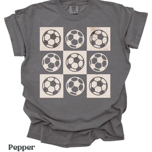 May include: A dark gray t-shirt with a white checkered pattern featuring nine soccer balls.