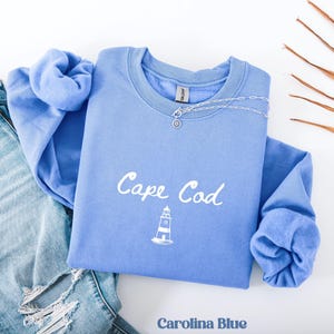 Può includere: Una felpa blu Carolina con la scritta "Cape Cod" in bianco e una grafica di un faro bianco. Una collana d'argento con un ciondolo è drappeggiata sulla felpa. La felpa è piegata sopra un paio di jeans blu.