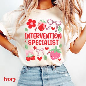 Puede incluir: Camiseta de color marfil con la frase "INTERVENTION SPECIALIST" en rojo. El diseño incluye gráficos de fresas y flores en rosa y rojo. La palabra "Ivory" está impresa en la parte inferior de la camiseta.