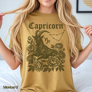 May include: Mustard-colored t-shirt featuring a Capricorn zodiac design. The graphic includes a goat, floral elements, the word "Capricorn", and the Capricorn constellation. The shirt is a solid color.