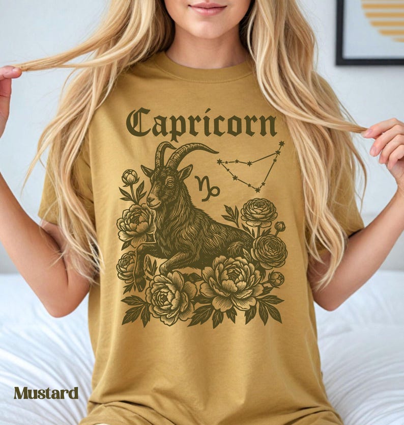 May include: Mustard-colored t-shirt featuring a Capricorn zodiac design. The graphic includes a goat, floral elements, the word "Capricorn", and the Capricorn constellation. The shirt is a solid color.