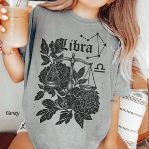 Op de afbeelding: Grijze t-shirt met een Weegschaal sterrenbeeld ontwerp. Het ontwerp bevat het woord "Libra", een weegschaal, een sterrenbeeld en bloemmotieven. De t-shirt is gemaakt van een zacht, comfortabel materiaal.