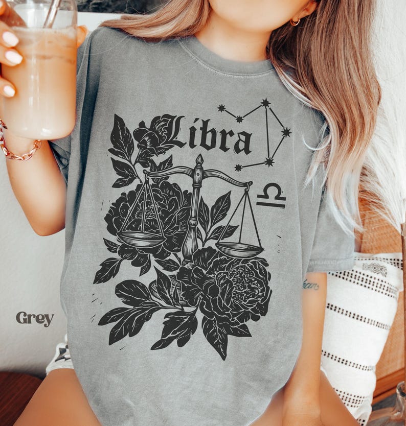 Op de afbeelding: Grijze t-shirt met een Weegschaal sterrenbeeld ontwerp. Het ontwerp bevat het woord "Libra", een weegschaal, een sterrenbeeld en bloemmotieven. De t-shirt is gemaakt van een zacht, comfortabel materiaal.