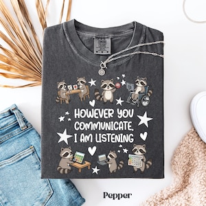 Könnte beinhalten: Dunkelgraues T-Shirt mit einem Waschbär-Motiv und dem Text "However You Communicate, I Am Listening". Das Design zeigt Waschbären, die verschiedene Kommunikationsmethoden verwenden. Das Shirt besteht aus einem weichen, bequemen Material.