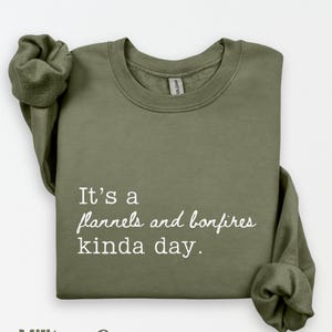 Puede incluir: Sudadera verde militar con el texto "It's a flannels and bonfires kinda day." La sudadera est&aacute; doblada con las mangas metidas. El texto "Military Green" est&aacute; en la parte inferior.