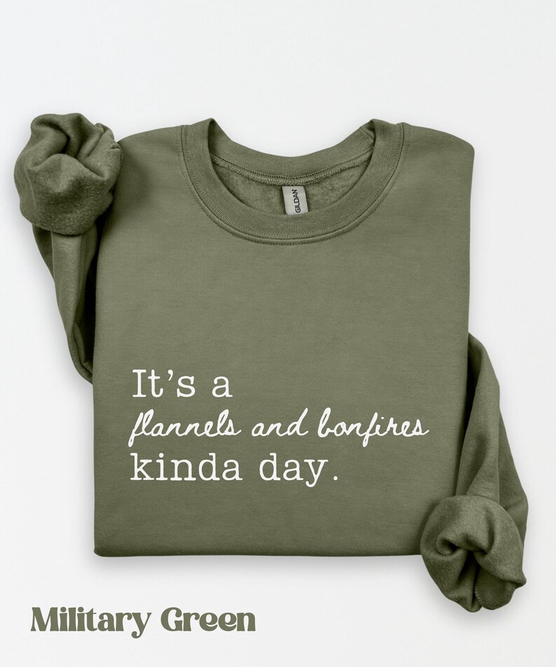 Puede incluir: Sudadera verde militar con el texto "It's a flannels and bonfires kinda day." La sudadera est&aacute; doblada con las mangas metidas. El texto "Military Green" est&aacute; en la parte inferior.
