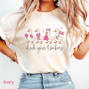 Pu&ograve; includere: T-shirt color avorio con un design di anatre dei cartoni animati. Le anatre indossano guantoni da boxe rosa, sciarpe e un fiocco. Il testo "check your honkers" &egrave; sotto. L'ultima anatra tiene una bandiera con "HOPE" e un nastro rosa.