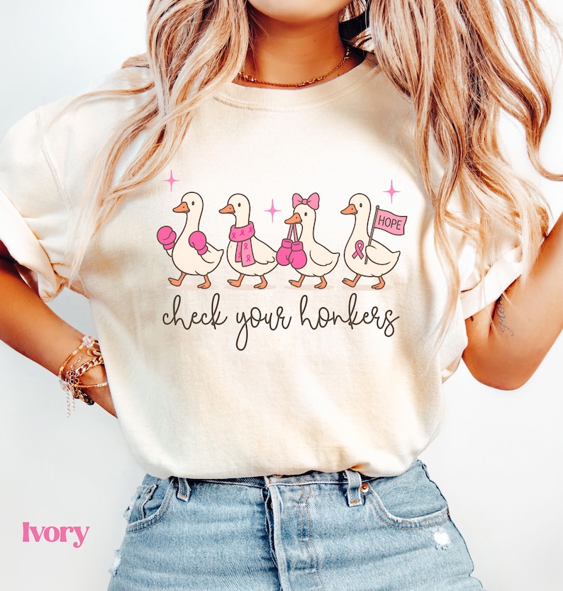 Pu&ograve; includere: T-shirt color avorio con un design di anatre dei cartoni animati. Le anatre indossano guantoni da boxe rosa, sciarpe e un fiocco. Il testo "check your honkers" &egrave; sotto. L'ultima anatra tiene una bandiera con "HOPE" e un nastro rosa.
