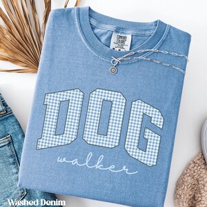 Könnte beinhalten: Ein T-Shirt mit Rundhalsausschnitt in verwaschenem Denim-Blau mit dem Wort DOG in großen Blockbuchstaben in einem hellblau-weißen Vichy-Muster. Unter den Buchstaben steht das Wort walker in weißer Schreibschrift. Eine silberne Halskette.